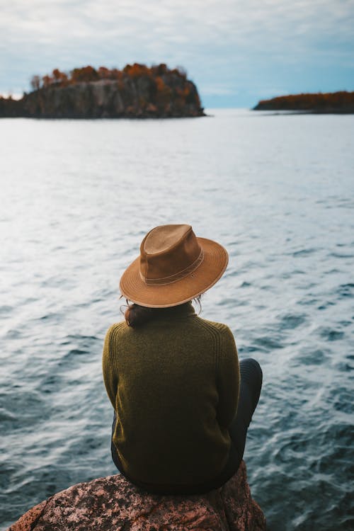 Back View of a Person in Brown Cowboy Hat and Green Sweatshirt Sitting on a Cliff Overlooking the Seascape Scenery