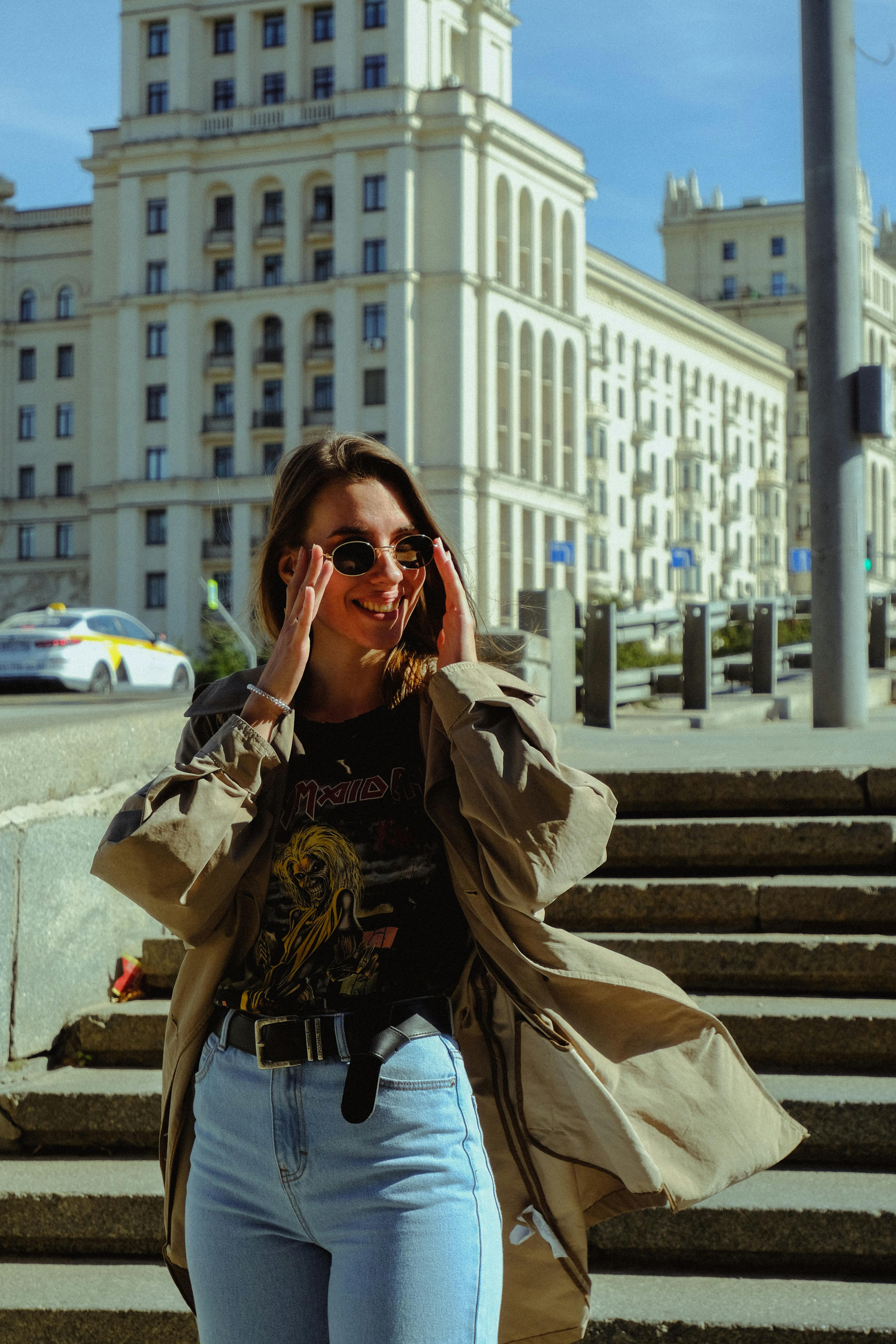 Young smiling female in stylish apparel touching sunglasses near staircase while looking away in town