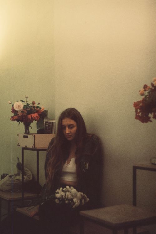 Free Pensive female with long hair looking down while sitting in room on chair with white flowers near wall with stands with bouquets Stock Photo