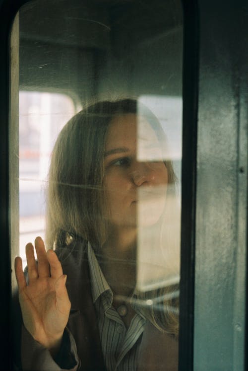 Thoughtful female looking away through window with hand on glass while standing in vestibule of train in daylight during journey