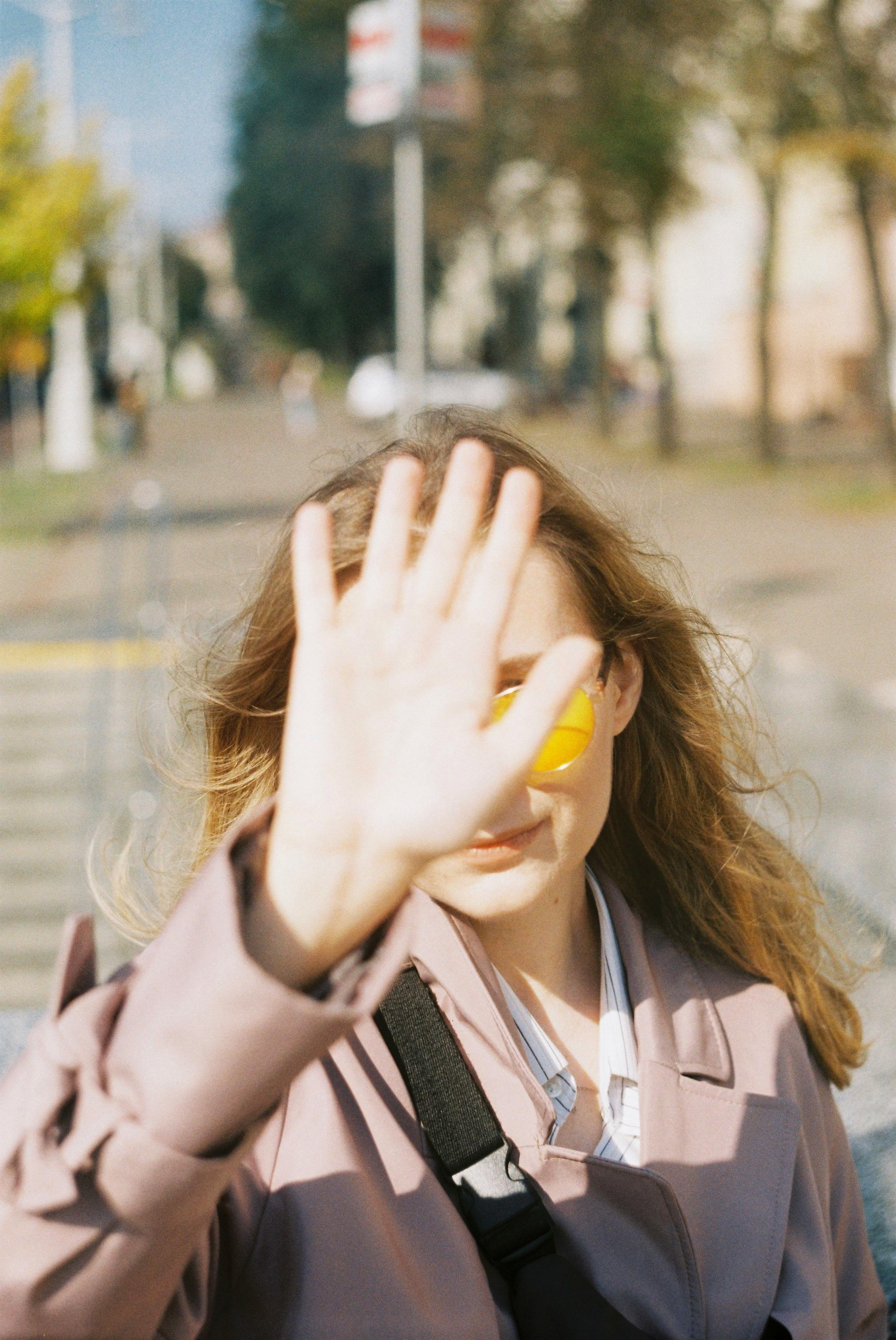 Stylish anonymous female with long hair showing stop gesture sign with palm and covering face while standing near crosswalk on blurred street