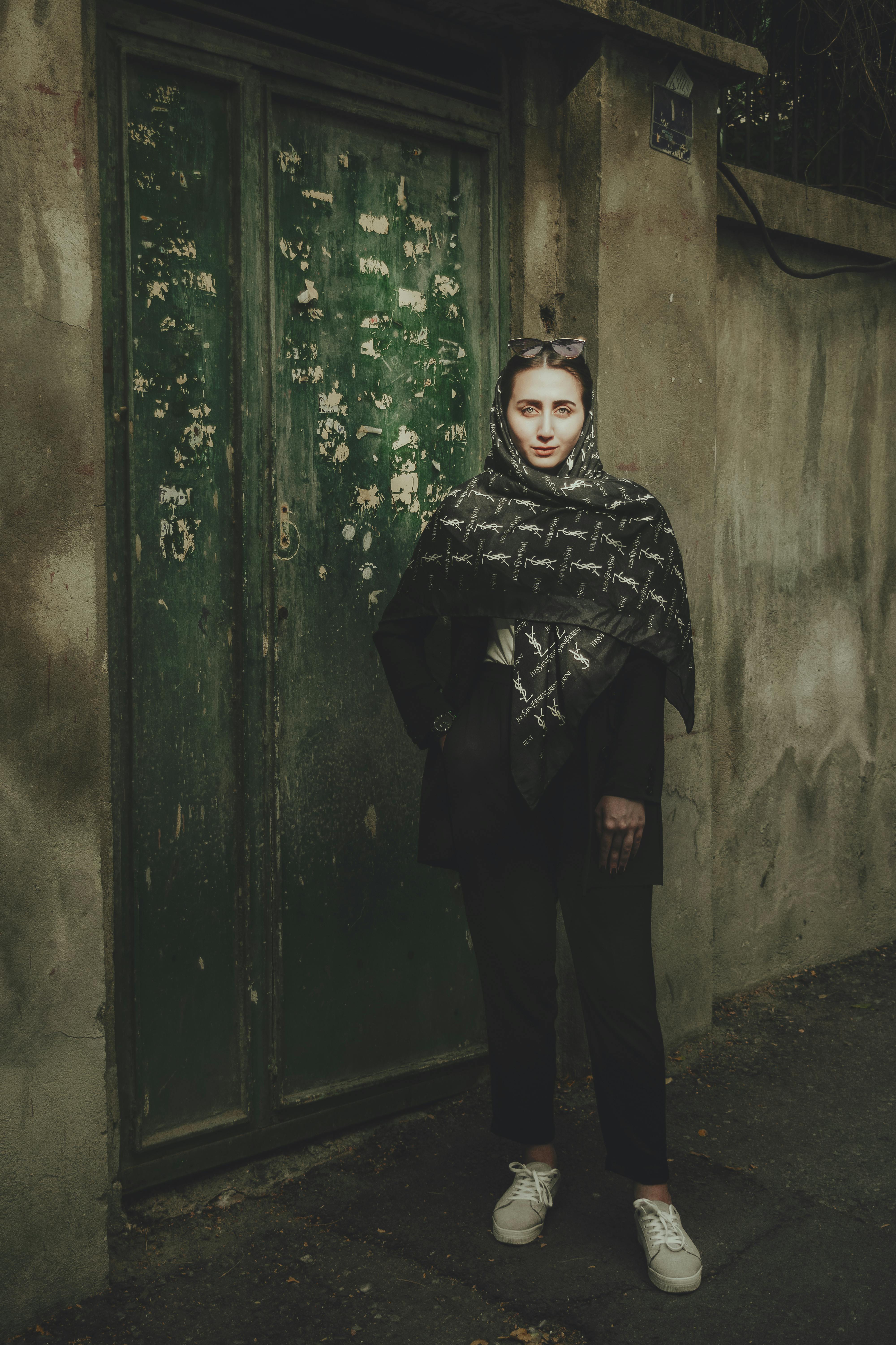 Young woman in scarf stands confidently by weathered green door in urban setting.