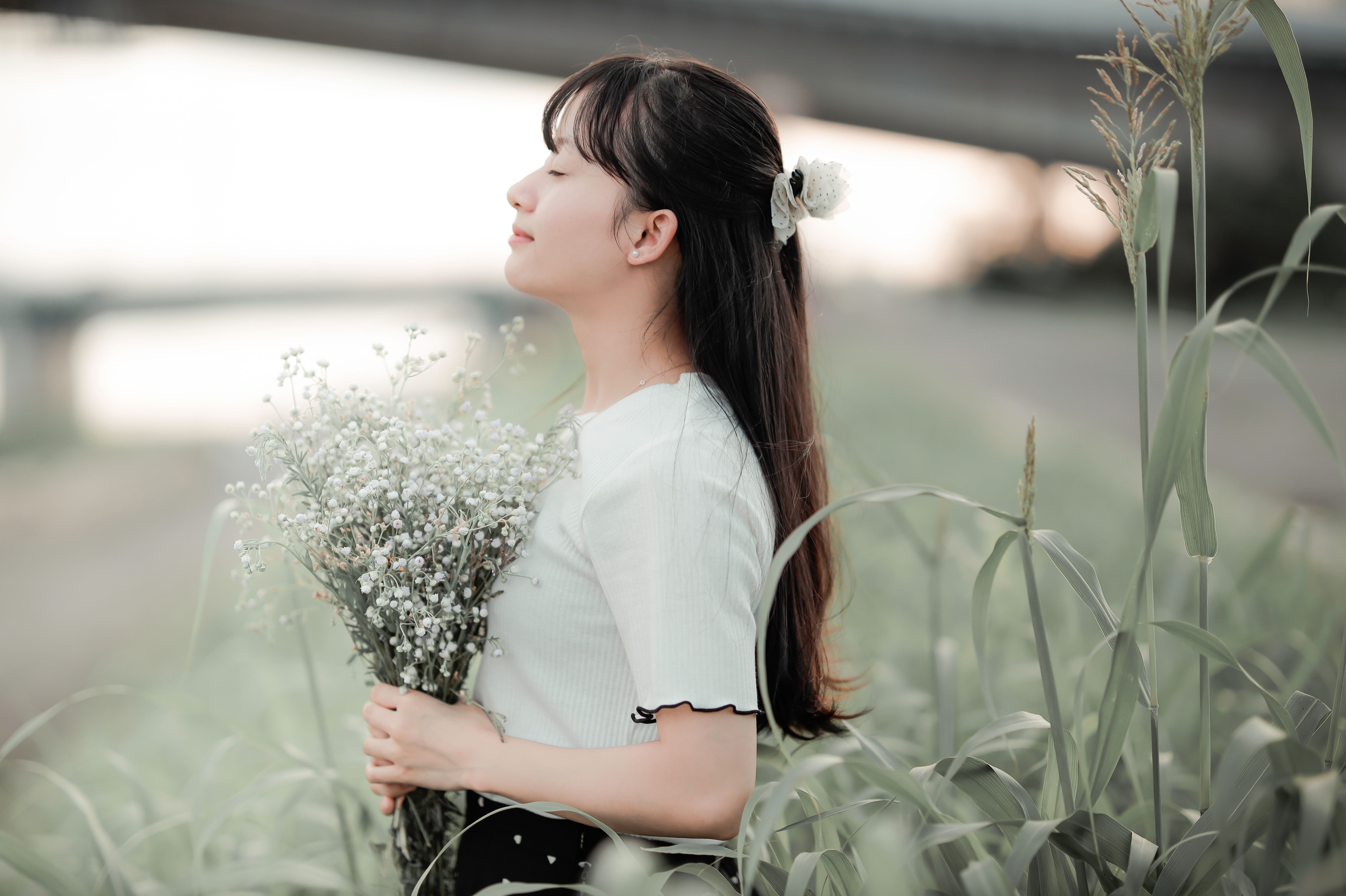 A woman stands in a grassy field holding a bouquet of wildflowers, eyes closed, feeling serene.