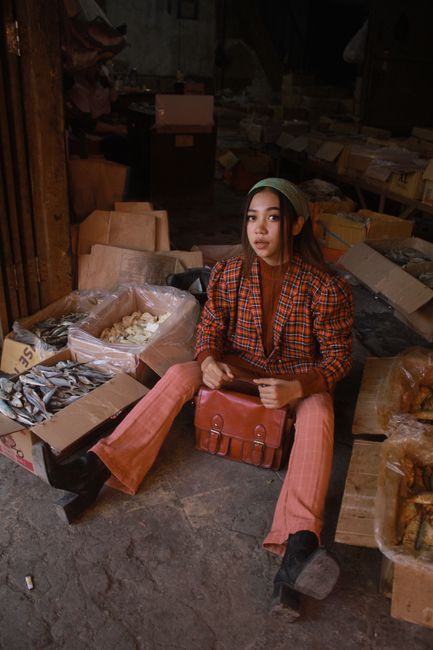From above of young ethnic female in trendy outfit and headband sitting on ground among boxes with various products in local market