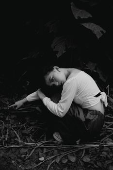Black and white of charming female in stylish clothes sitting on ground in nature in daylight