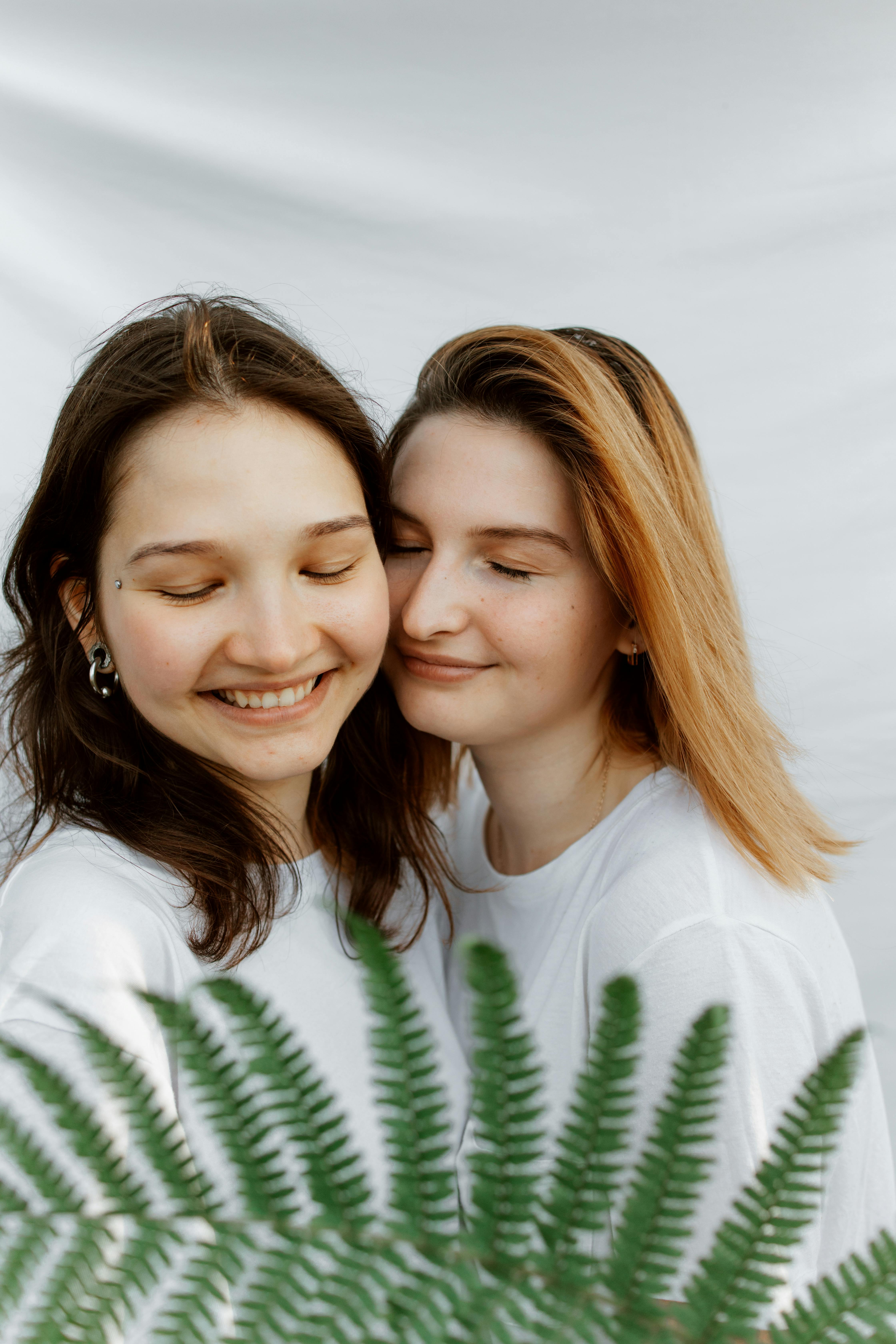 Two smiling women with closed eyes embracing behind lush ferns.