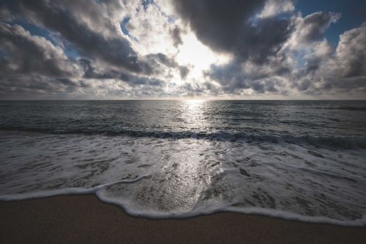 Picturesque view of ocean foamy waves on sandy beach under clouds at sundown
