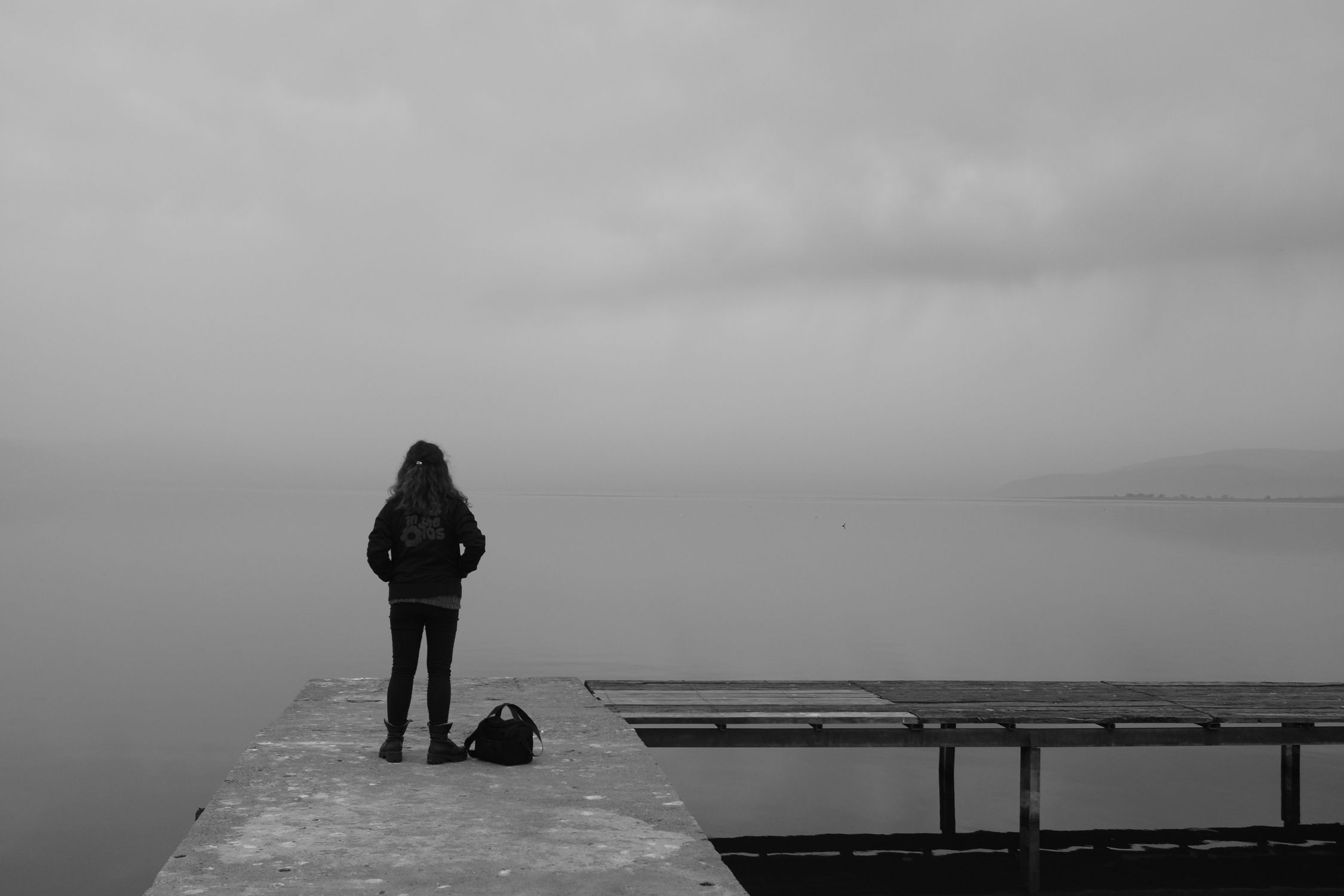 Foto monocromática de uma mulher em pé em um píer nublado com vista para um lago tranquilo em İznik, na Turquia.