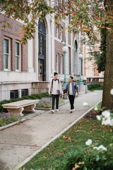 Two friends walking together on a college campus sidewalk, enjoying a casual conversation.