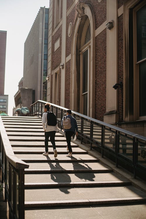 Back view of anonymous friends in casual clothes with backpacks walking upstairs along building in city in sunny day