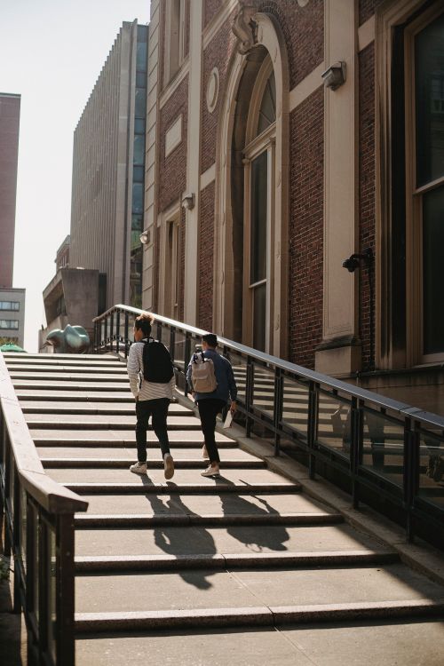 Free Back view of anonymous friends in casual clothes with backpacks walking upstairs along building in city in sunny day Stock Photo