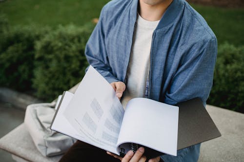 Free Crop anonymous student in casual clothes sitting on bench in green park and learn project with graphs in daytime Stock Photo