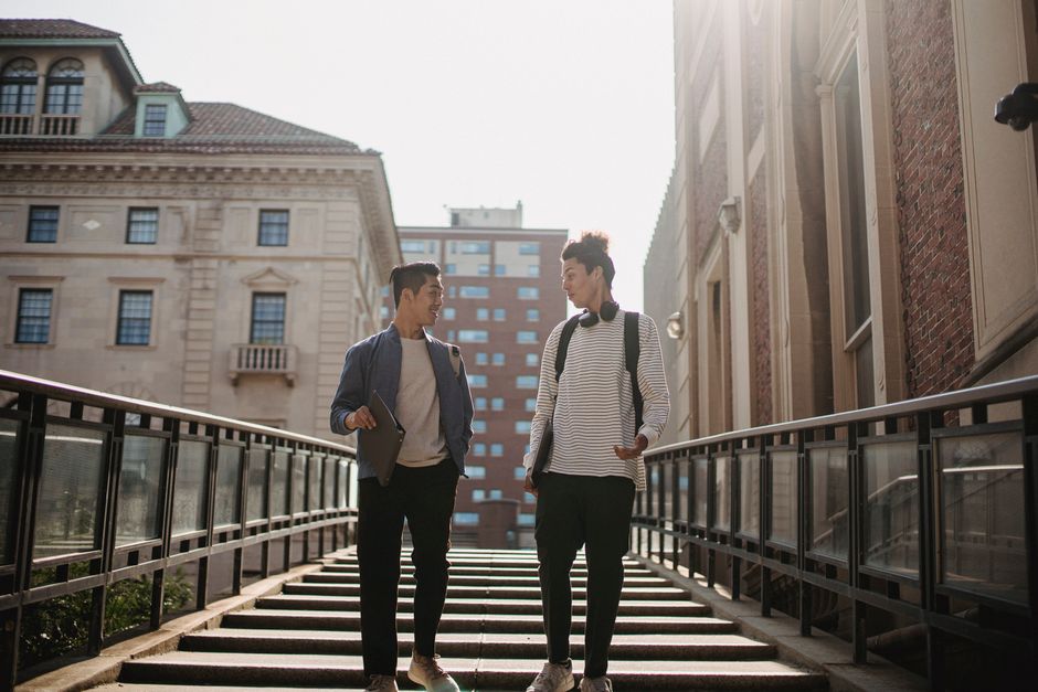 Low angle of positive multiracial male students with backpacks and documents strolling on staircase and speaking to each other Low angle of positive multiracial male students with backpacks and documents strolling on staircase and speaking to each other