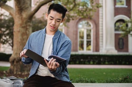 Free Concentrated Asian male student sitting on bench and reading information on papers in folder for homework Stock Photo