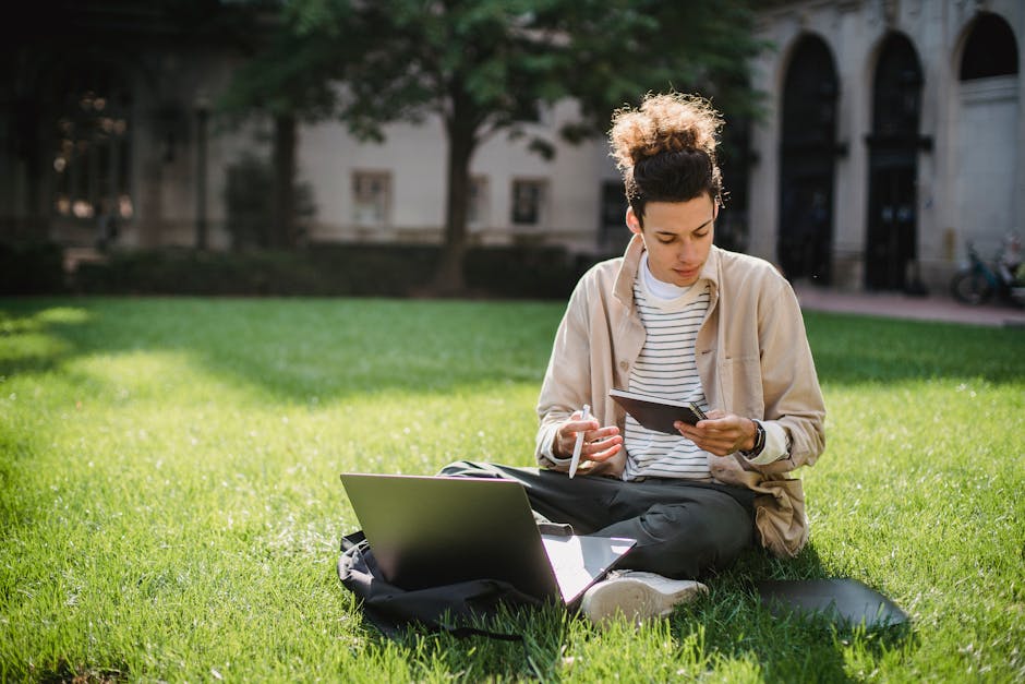 Concentrated student sitting on grass with notepad and laptop while working on homework assignment in park