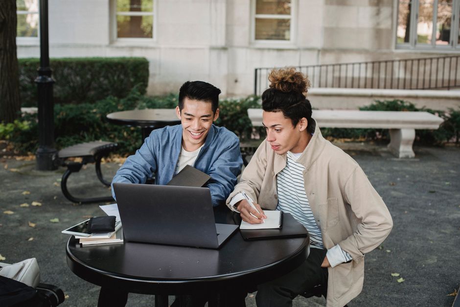 Smiling multiracial male classmates sitting at table and browsing netbook while taking notes in notebook Smiling multiracial male classmates sitting at table and browsing netbook while taking notes in notebook