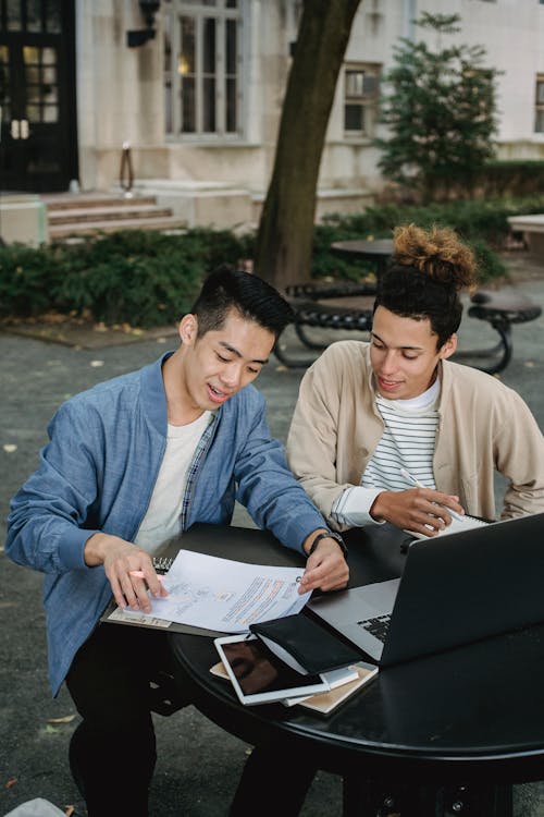 Focused young male students in casual wear sitting at table with laptop and copybooks in university campus and preparing for exams together