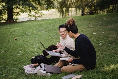 Side view full length smiling ethnic male students in casual outfits resting on grassy lawn with laptop and textbooks while preparing for exams together in green park