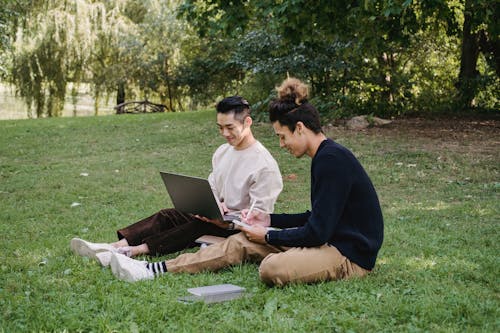 Full body smiling young ethnic male students in smart casual outfits using modern netbook and taking notes in copybooks while working on assignment on park lawn