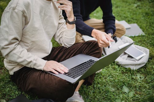 Free Crop anonymous male student in smart casual wear browsing netbook and discussing university project while sitting on grass in lush garden Stock Photo