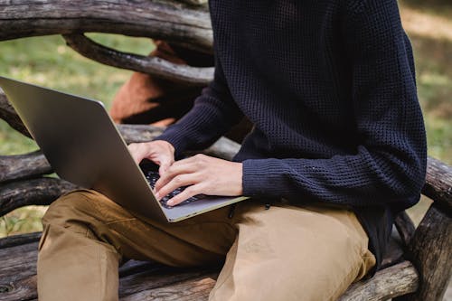 Free Crop anonymous male in casual clothes working on modern netbook while sitting on creative wooden bench in park Stock Photo