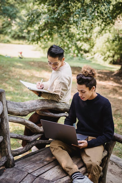 Happy young ethnic male students in casual outfits browsing modern laptop and taking notes in notebook while studying together in sunny park