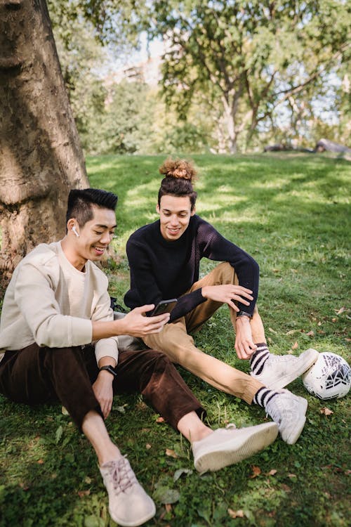 Free Smiling young ethnic males in casual clothes watching funny video on modern mobile phone while resting together on grass meadow in park Stock Photo