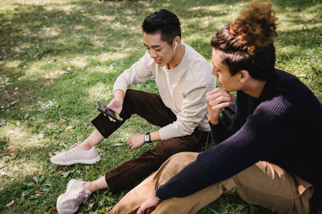 From above content young ethnic male friends wearing casual outfits surfing internet on modern mobile phone and sitting on grass in sunny park