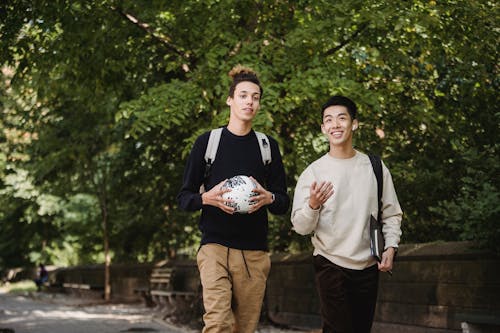 Happy smiling ethnic male friends with backpacks and soccer ball strolling together in lush park and chatting
