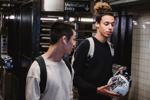 Positive young male friends in casual sweaters carrying backpacks and soccer ball chatting and walking in modern underground passage