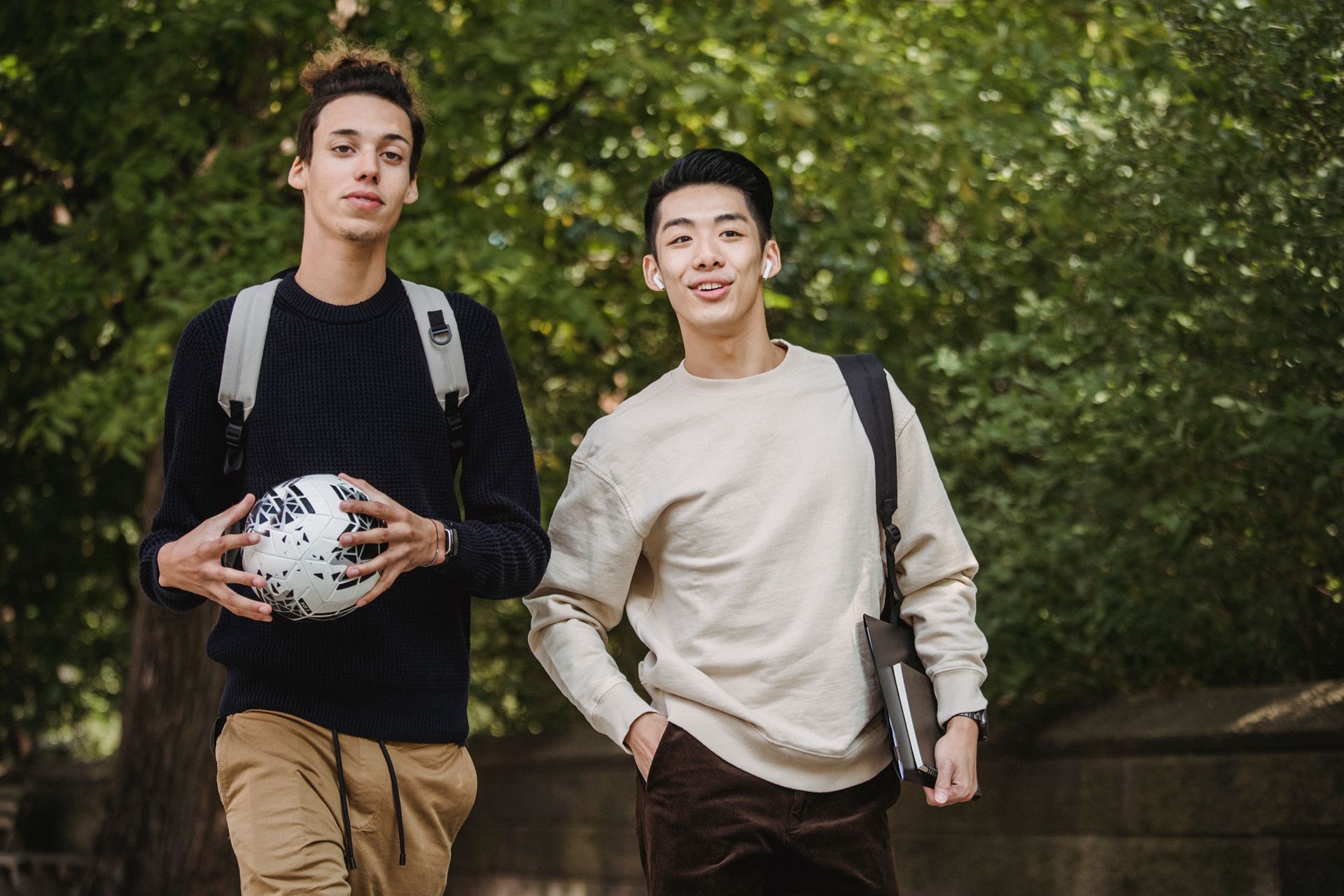 Two young men walking on a park path, one carrying a soccer ball and the other carrying a textbook