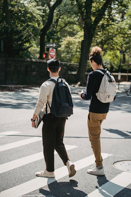 Anonymous young men crossing road near green trees in sunlight