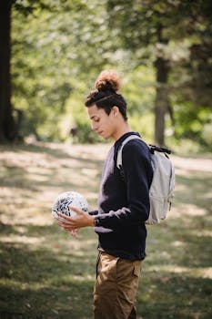 阳光明媚的日子里，一位留着时尚发型的少数族裔男性青少年身着休闲服，背着背包，手拿足球，侧身沉思