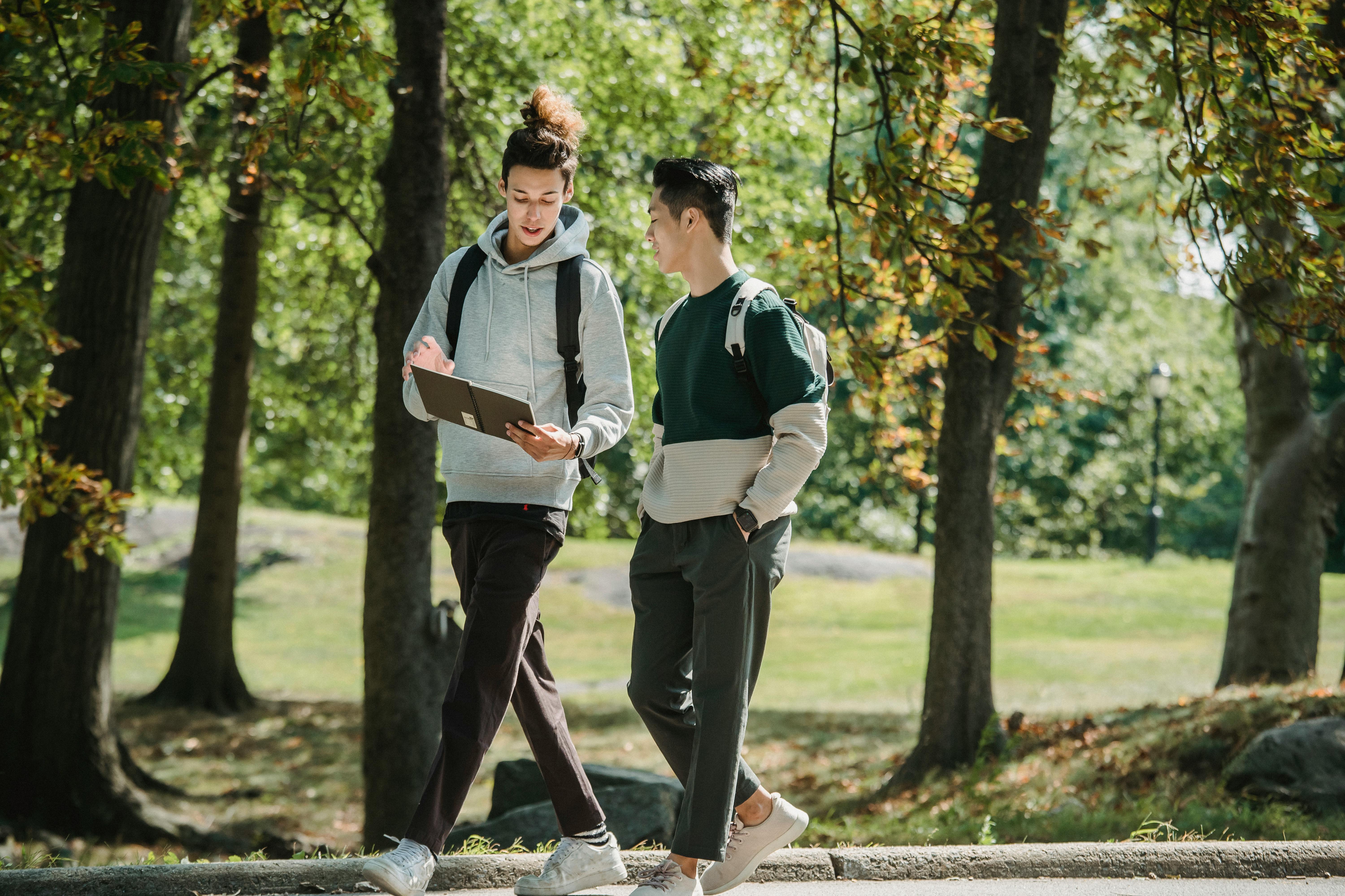 concentrated young diverse male students strolling in park and reading notepad