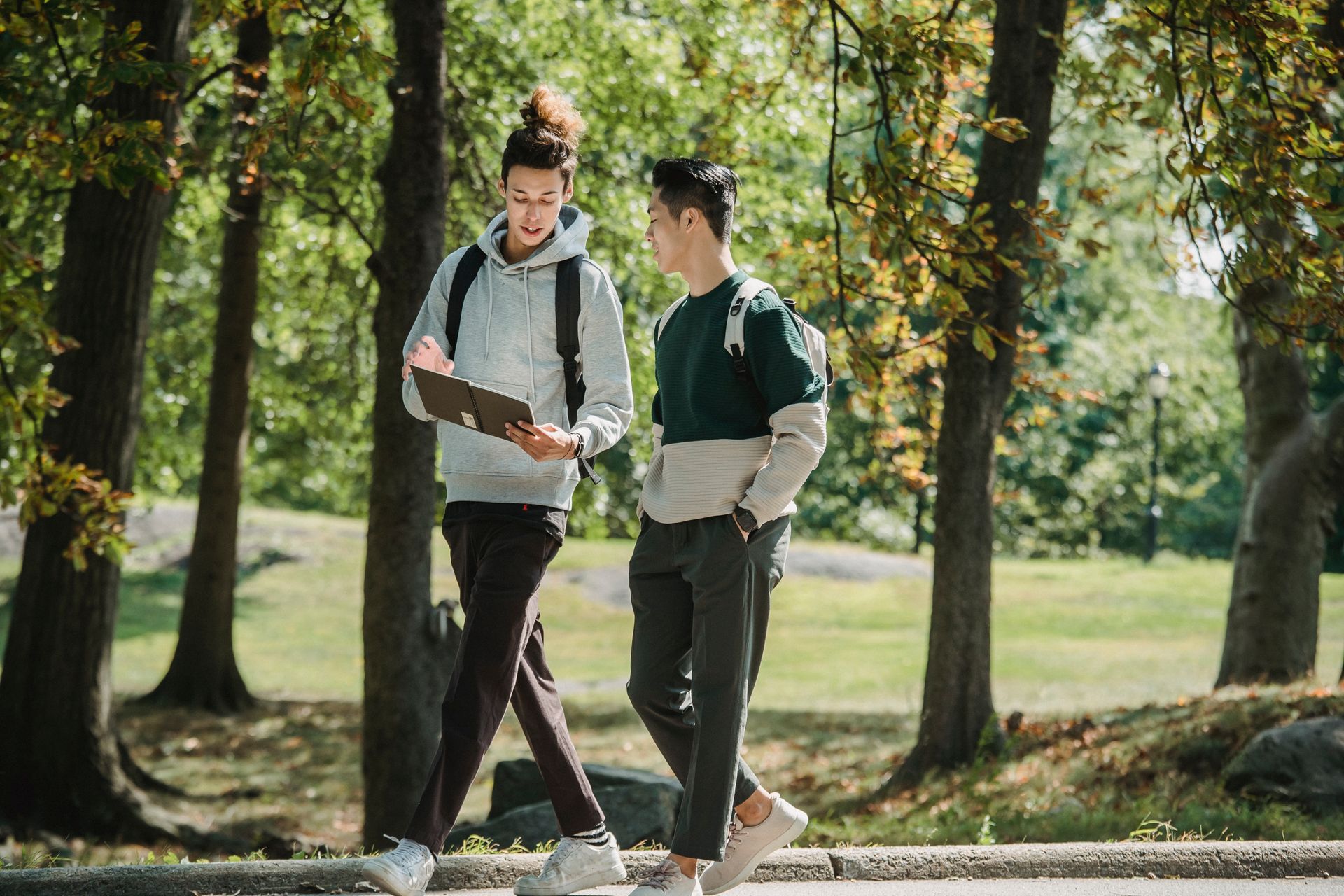 Concentrated young diverse male students strolling in park and reading notepad