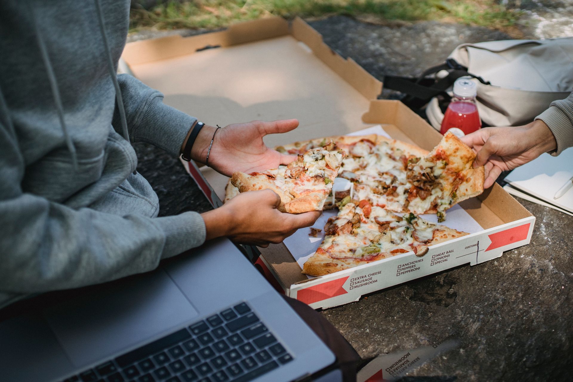 Two friends enjoying pizza outdoors with a laptop, embodying leisure and friendship.