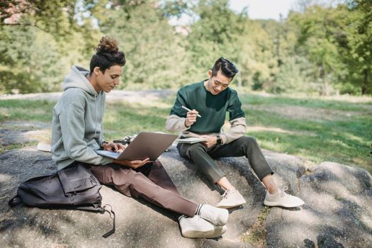Full length young men friends in casual outfit sitting on big boulder with computer while writing notes with pen in notepad in summer sunny day with trees on background