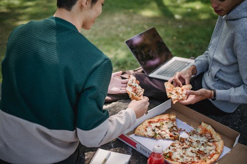 High angle of crop faceless young male friends spending time on boulder near grass with computer and pizza and with notepad and pen in summer sunny day