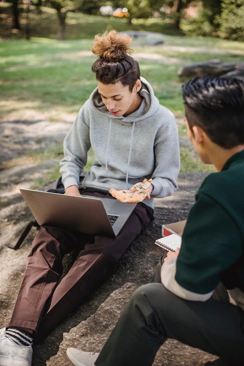 Focused male learners using laptop and having pizza during homework preparation in park