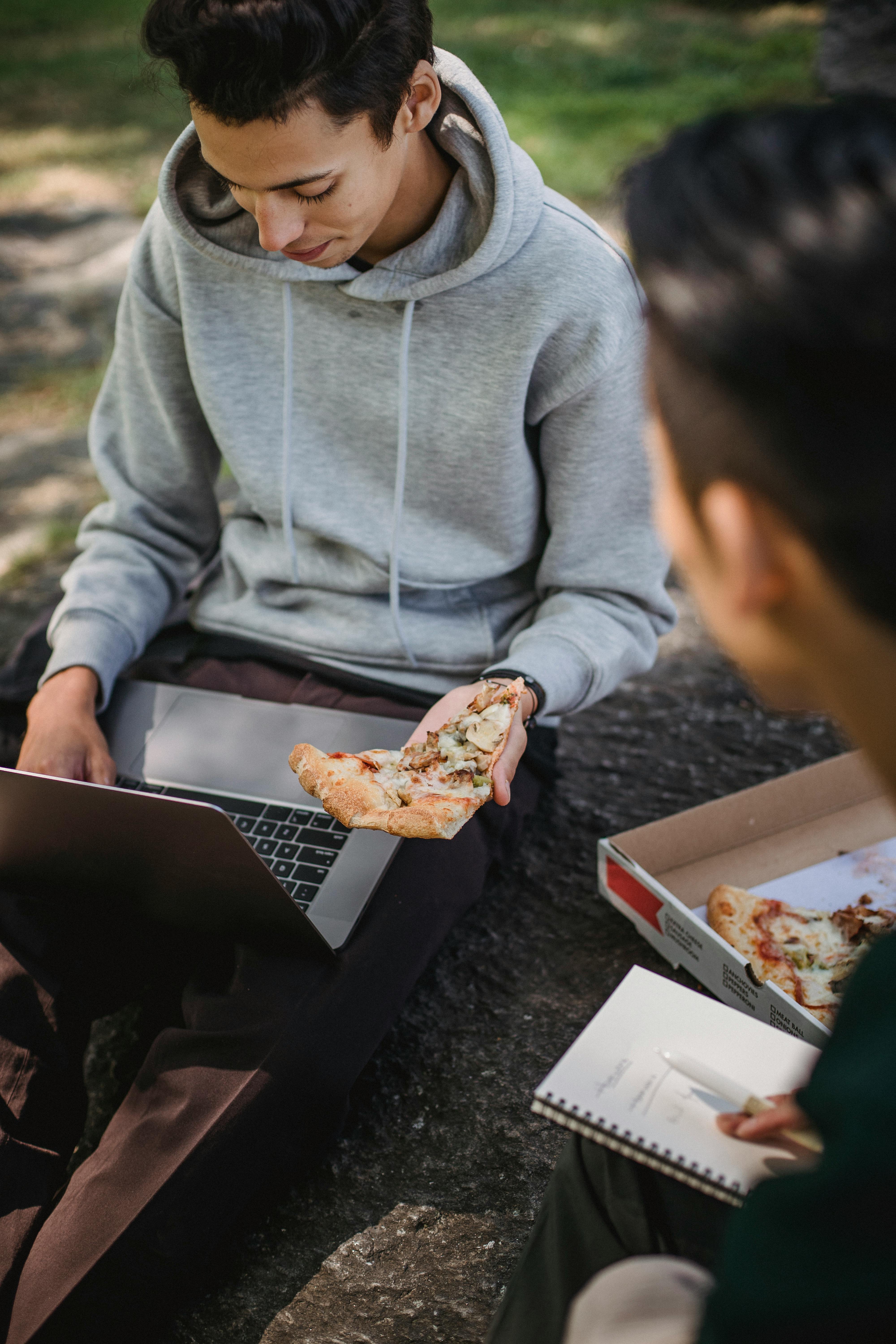 students using laptop and eating pizza in park