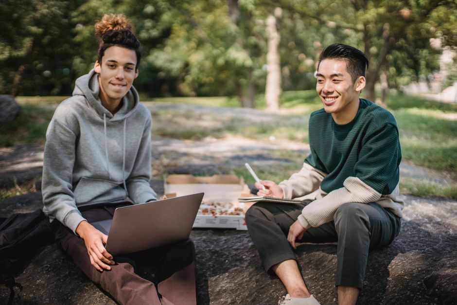 Positive multiracial male classmates sitting on stone with laptop and pizza while doing homework task together Positive multiracial male classmates sitting on stone with laptop and pizza while doing homework task together