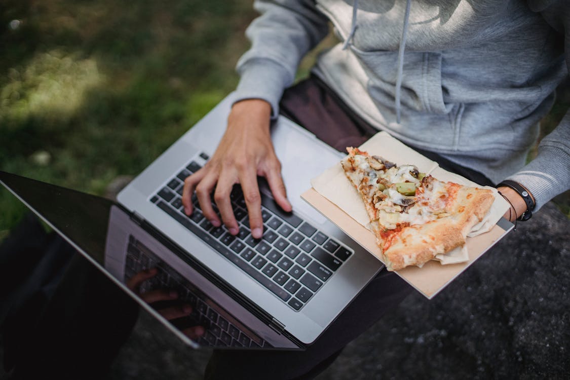 From above of crop anonymous learner using laptop for studies and eating pizza in park