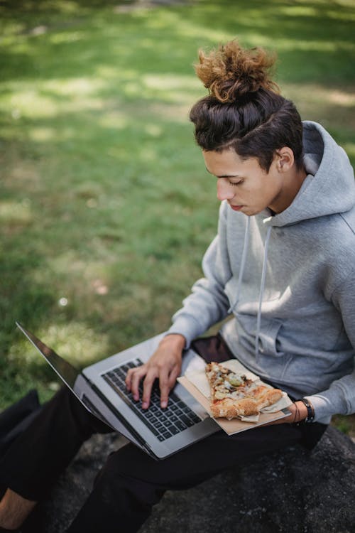 Serious male student doing homework with laptop and pizza