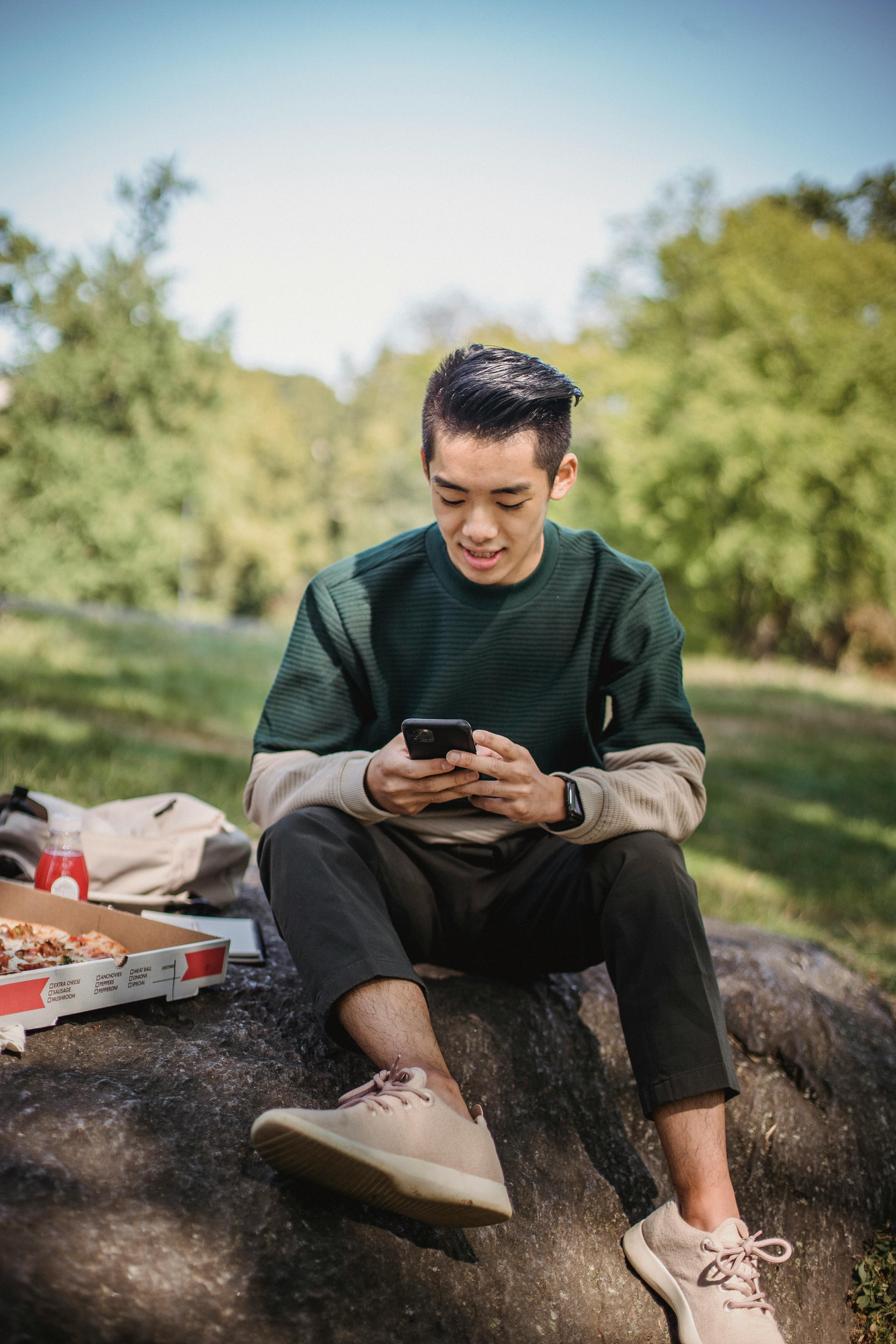 asian man using smartphone sitting on stone in park