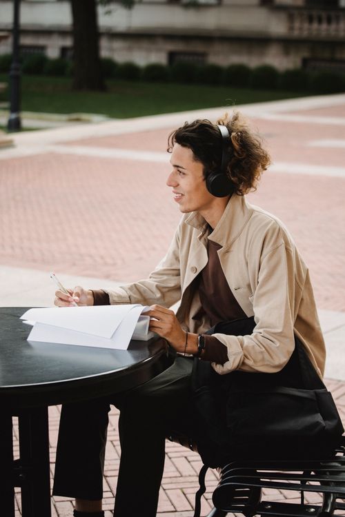 Free Side view of positive student smiling while sitting on bench and doing homework assignment while listening to music in wireless headphones Stock Photo