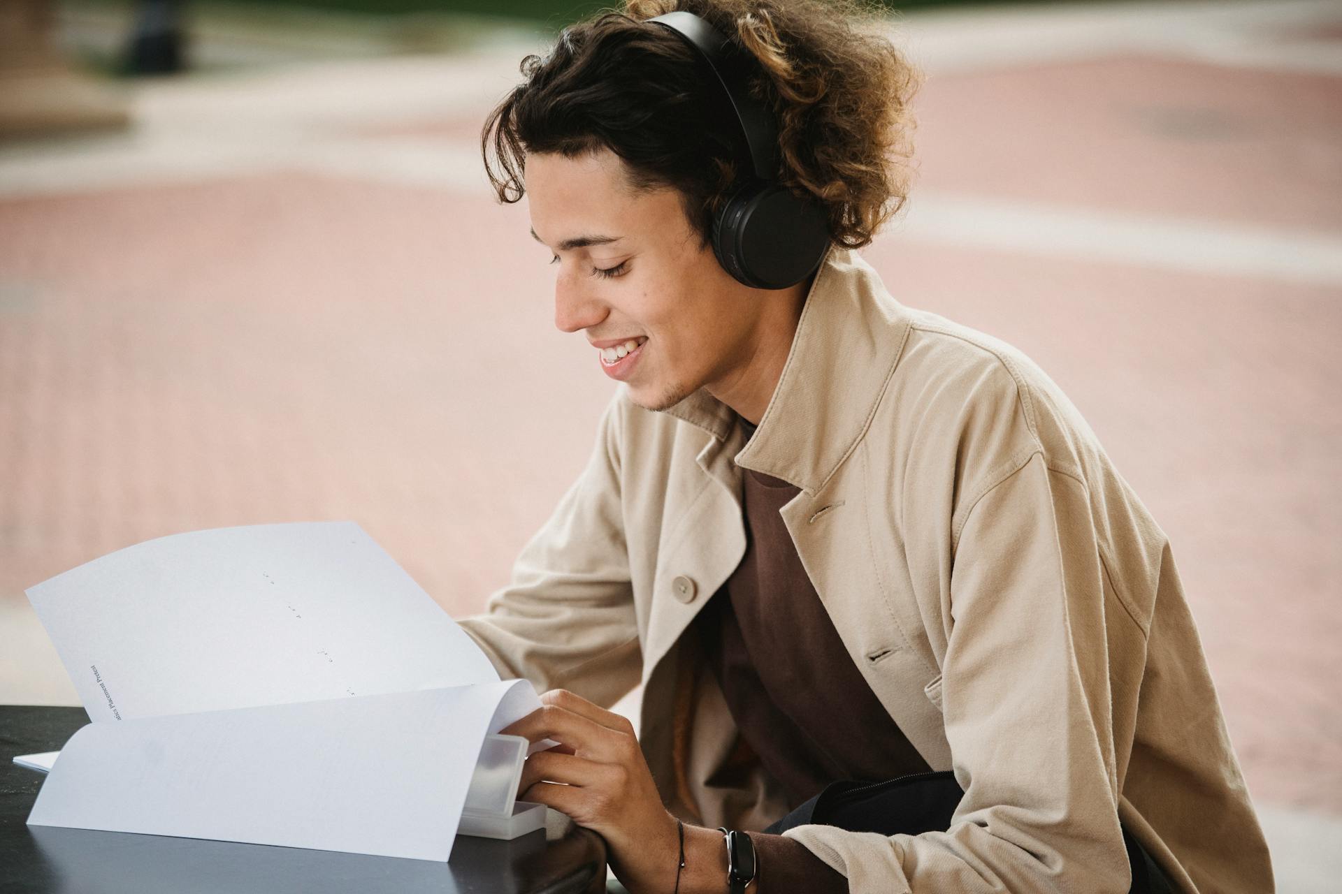 A young male student enjoying studying outdoors with headphones, focused and smiling.