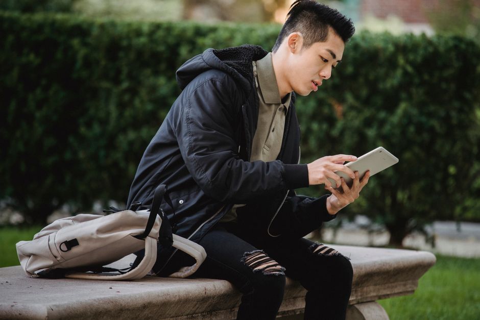Side view of serious Asian male learner in casual clothes with backpack surfing tablet while sitting in park Side view of serious Asian male learner in casual clothes with backpack surfing tablet while sitting in park