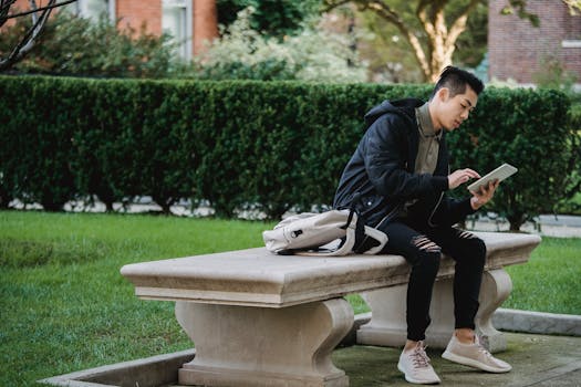 Full body of young focused Asian male in casual wear sitting on bench in park and browsing internet on tablet