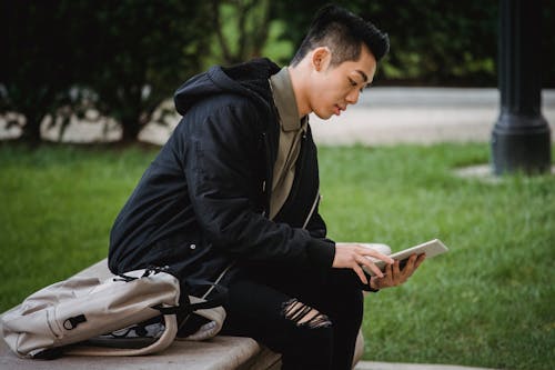 Side view of focused young Asian male student in casual clothes sitting on bench in park with backpack and browsing internet on tablet