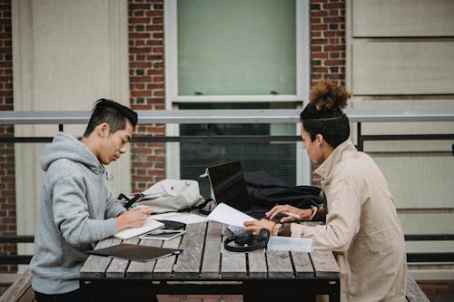 Side view of young concentrated multiracial male students in casual outfits sitting at table with gadgets and copybooks while working on homework